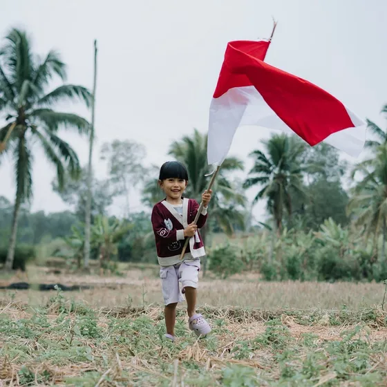 A young girl waving the Indonesian flag in a rice field lined with palm trees
